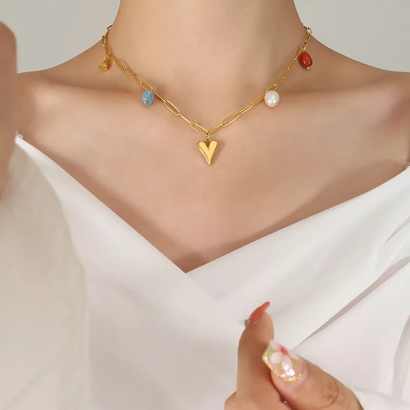 Une personne portant un collier en or avec un pendentif en forme de cœur et des breloques colorées, vêtue d'un haut blanc décolleté, tenant ses mains près de sa poitrine.