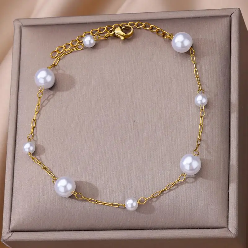 Un bracelet en chaîne en or avec six perles blanches régulièrement espacées, présentées sur une surface beige coussinée.