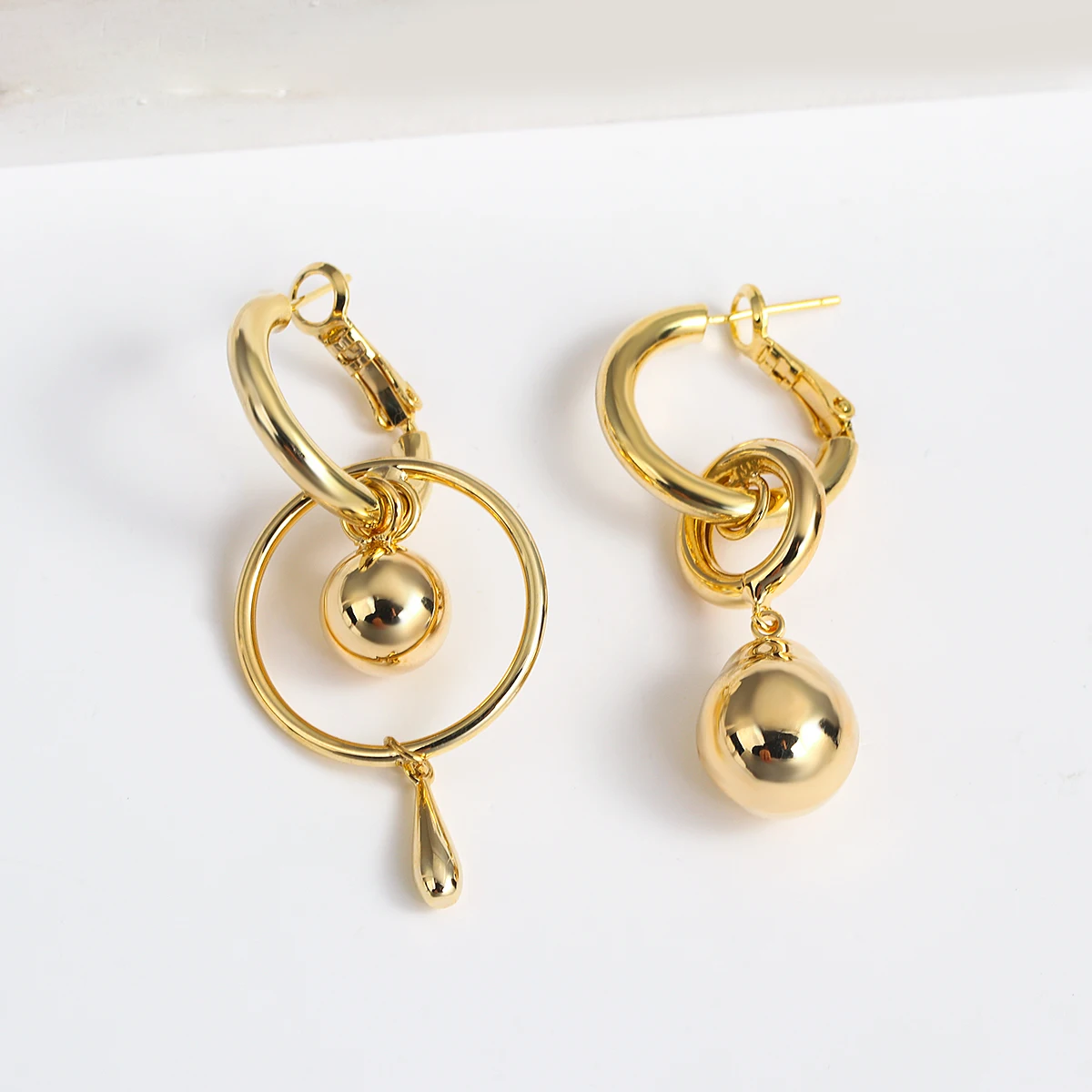Une paire de boucles d'oreilles dépareillées dorées, chacune avec une perle ronde en or et des gouttes décoratives distinctes sur des anneaux asymétriques.