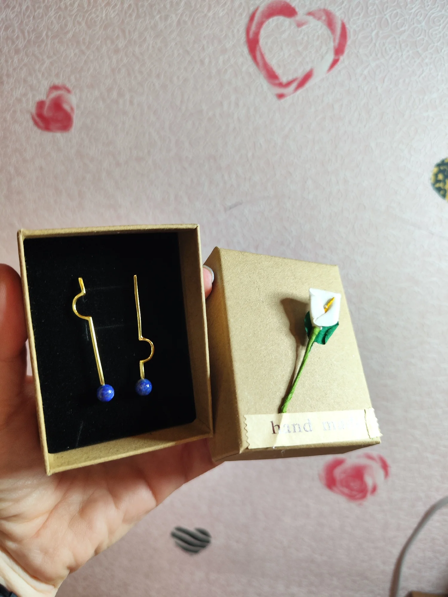 Une main tenant une boîte ouverte avec une paire de Boucles d'oreilles asymétriques avec perles bleues sur un coussin noir et une petite fleur décorative sur le couvercle de la boîte.