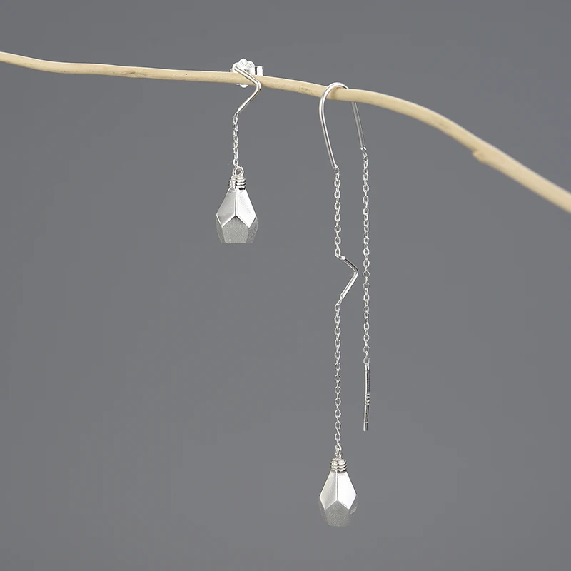Une paire de boucles d'oreilles pendantes géométriques argentées ROSY Boucles d'oreilles asymétriques goutte avec crochets de chaîne est suspendue à une fine branche de couleur claire sur un fond gris uni.