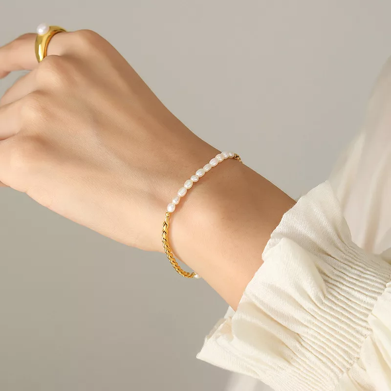 Une main portant un anneau d'or et deux bracelets, l'un en chaîne d'or et l'autre avec de petites perles blanches, sur un fond uni. La manche est blanche et légèrement froncée.