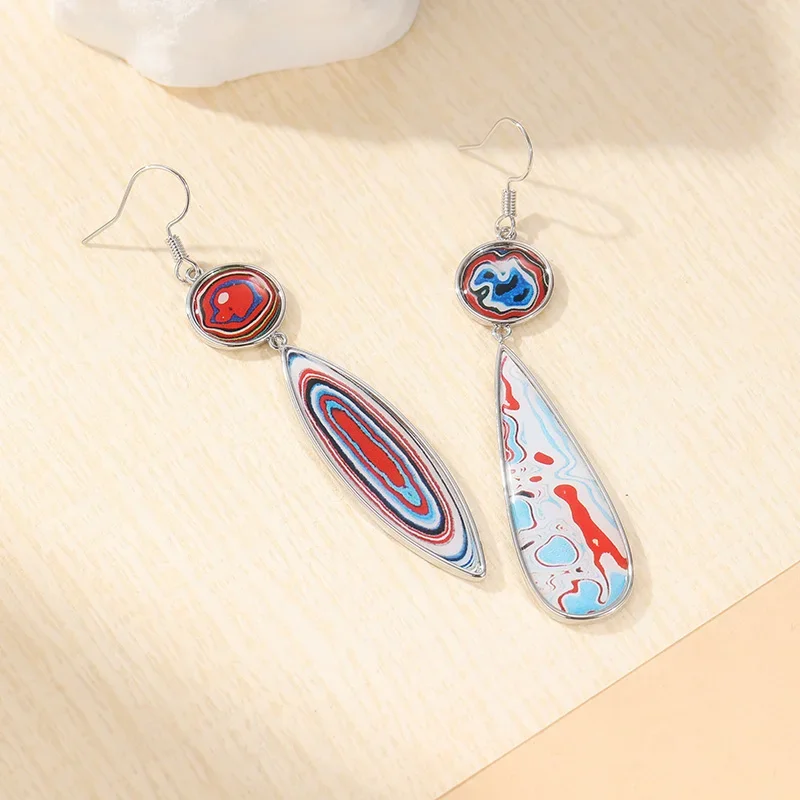 Paire de boucles d'oreilles dépareillées, chacune avec une forme ronde et une forme allongée en goutte d'eau, présentant des motifs marbrés rouges, bleus et blancs, présentés sur une surface en bois clair.
